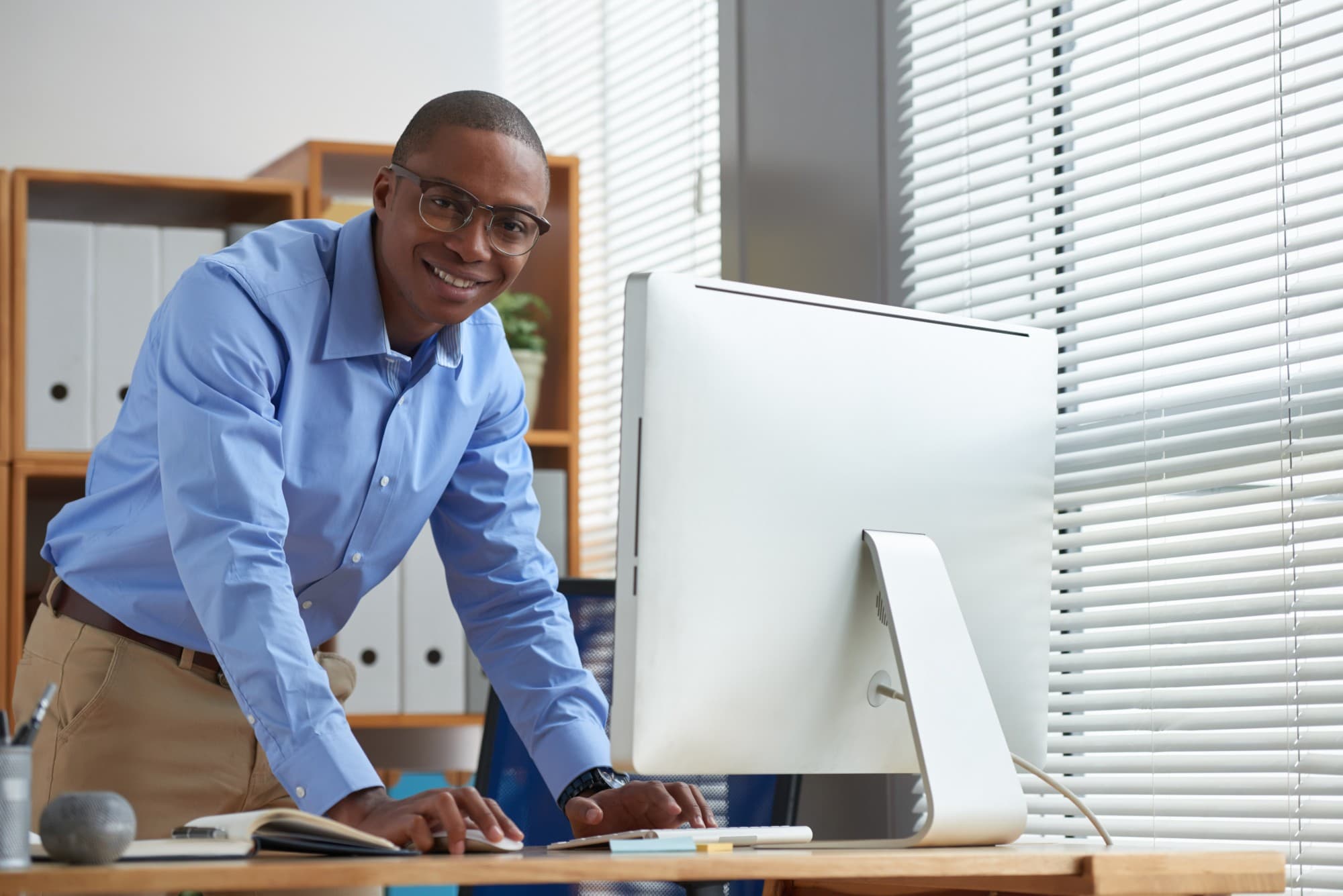 A smiling professional standing at a desk with a desktop computer in a bright office